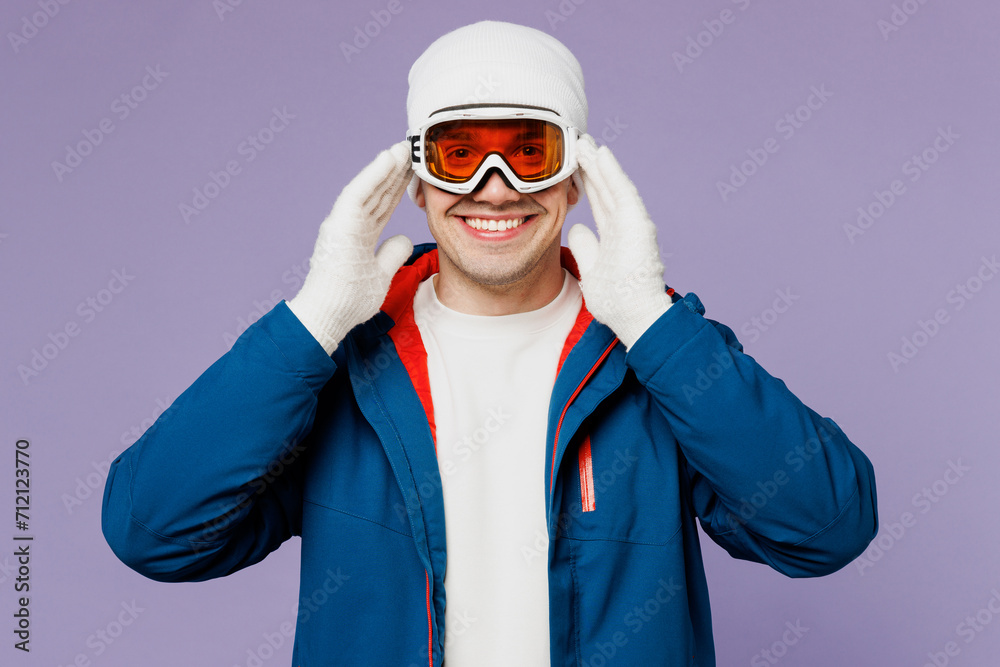 Skier smiling happy cheerful man wear warm blue windbreaker jacket touch ski goggles mask hat looking camera spend extreme weekend winter season in mountains isolated on plain light purple background. Skier smiling happy cheerful man wear warm blue windbreaker jacket touch ski goggles mask hat looking camera spend extreme weekend winter season in mountains isolated on plain light purple background.