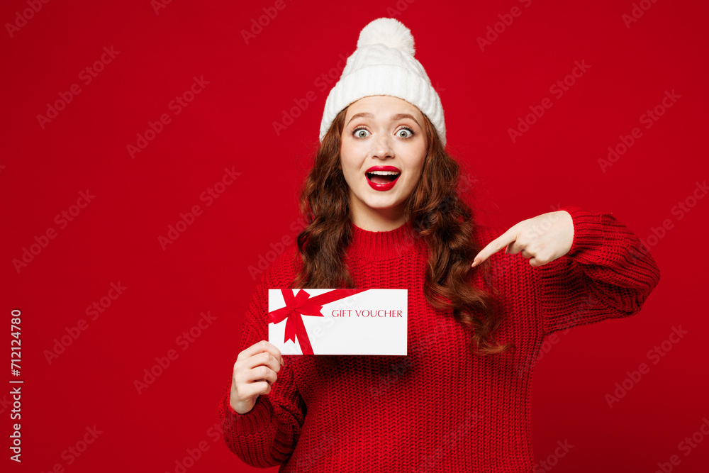 Young amazed woman wears knitted sweater white hat casual clothes hold point index finger on gift certificate coupon voucher card for store isolated on plain red color background. Lifestyle concept. Young amazed woman wears knitted sweater white hat casual clothes hold point index finger on gift certificate coupon voucher card for store isolated on plain red color background. Lifestyle concept.