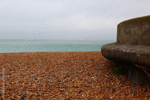 Empty pebble beach on England's south coast on an overcast, rainy, grey, summer day with cloudy skies, a rough see, and an empty cement curved bench. Beach with rocks and waves and ample copy space