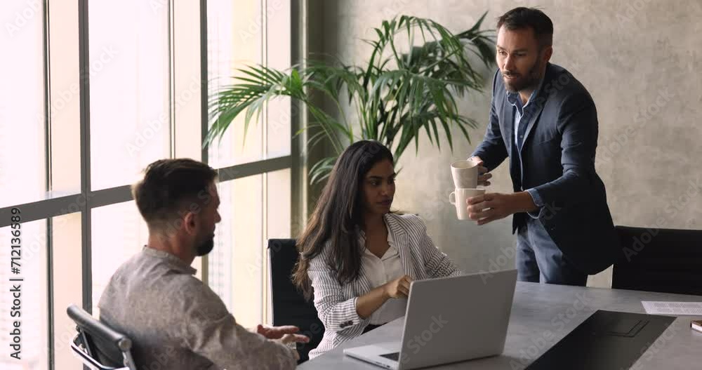 Middle-aged colleague in suit bringing tea cups, morning beverage to multiethnic teammates, team members having coffee break during workday gathered in modern conference room. Pause, friendly relation