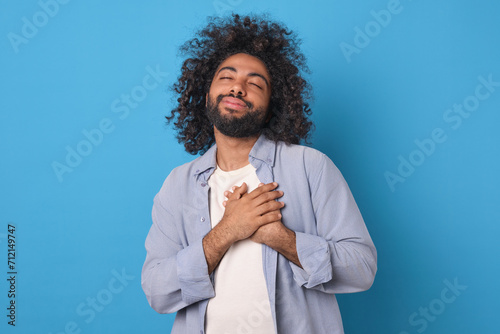 Young attractive happy Arabian man puts hands on heart and with eyes closed raises head showing gratitude or empathy and wishing well to people around posing on isolated blue background photo