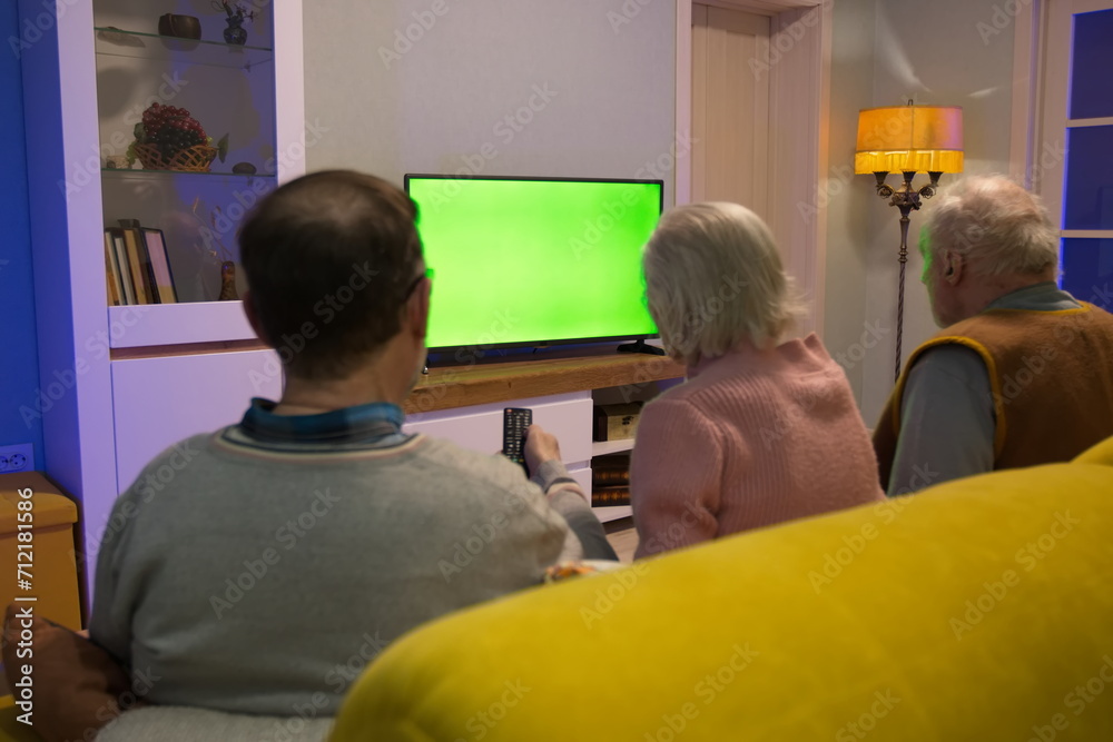 Family watching TV. Green screen. An adult man and his elderly parents are sitting at home on the couch and watching TV. A man holds a TV remote control in his hand. Family watching TV. Green screen. An adult man and his elderly parents are sitting at home on the couch and watching TV. A man holds a TV remote control in his hand.