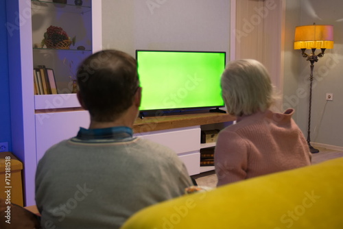 Family watching TV. Green screen. An adult man and his elderly mother are sitting at home on the couch and watching TV. In front of them is a green screen TV. 