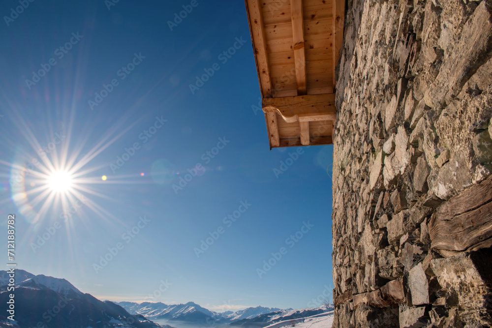 part of a stone cottage with snow-covered mountain views and sun shining in the sky