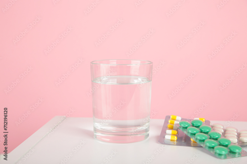 Pink alarm clock with glass of water and blister packages of pills on light table background.