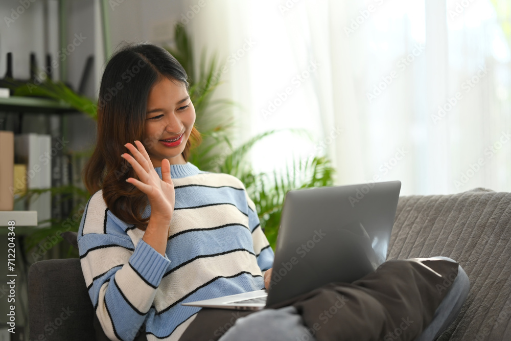 Beautiful young woman in sweater sitting on sofa at home and having video call via laptop Beautiful young woman in sweater sitting on sofa at home and having video call via laptop