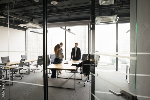 Successful multiethnic business team meeting for cooperation, brainstorming, networking in office co-working space, conference room, standing at large table, talking. Through glass wall, doorway view
