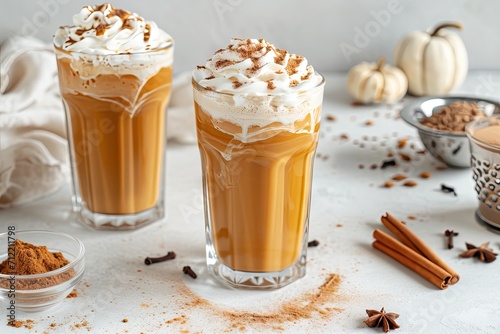 Two pumpkin spice lattes in tall glasses with whipped cream chocolate dust and spices nearby on a light grey table