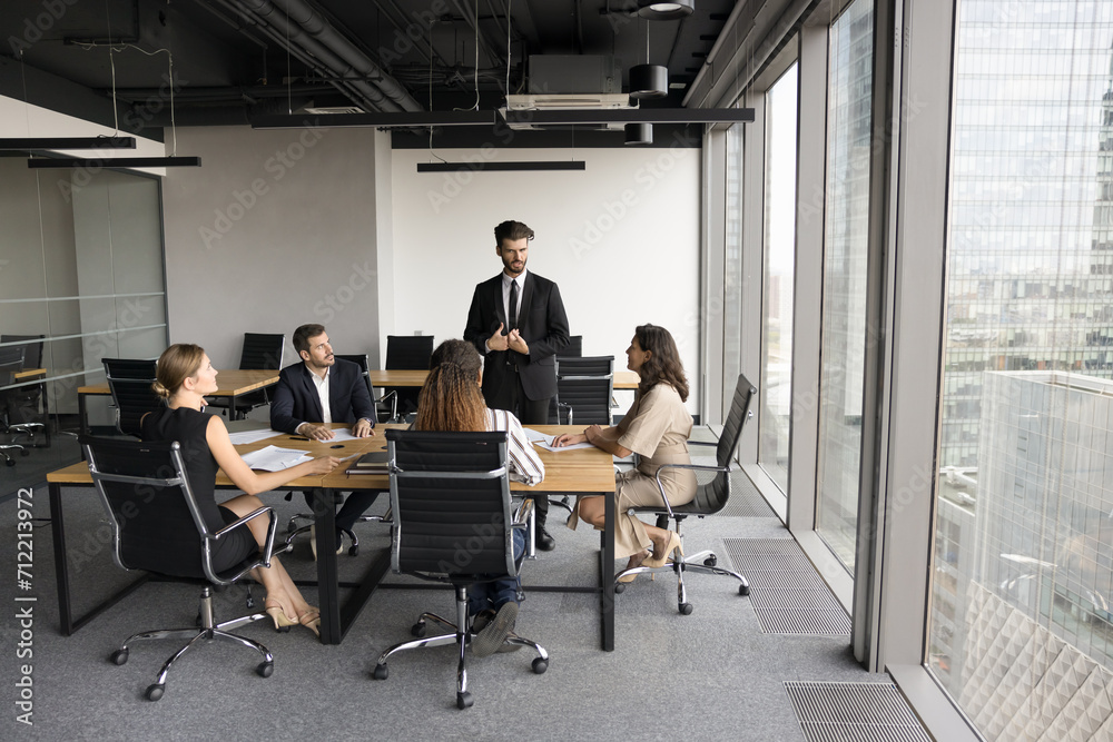 Serious business leader man in formal suit speaking to team on meeting in modern urban office space, standing at table, offering cooperation plan to diverse colleagues. Full length wide shot