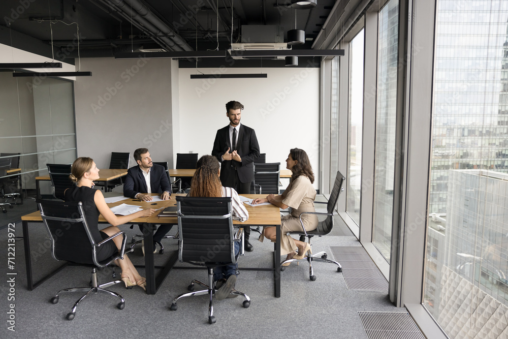 Serious business leader man in formal suit speaking to team on meeting in modern urban office space, standing at table, offering cooperation plan to diverse colleagues. Full length wide shot Serious business leader man in formal suit speaking to team on meeting in modern urban office space, standing at table, offering cooperation plan to diverse colleagues. Full length wide shot