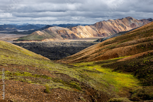 View at volcanic mountains with snow, green moss on hills and black lava on Icelandic highlands at Landmannalaugar, Iceland. Famous Laugavegur hiking trail with dramatic sky