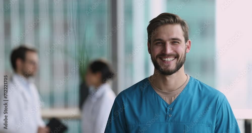 Serious young handsome doctor man in blue uniform shirt getting cheerful, happy, looking at camera with toothy smile, standing in hospital hall space with colleagues in background Serious young handsome doctor man in blue uniform shirt getting cheerful, happy, looking at camera with toothy smile, standing in hospital hall space with colleagues in background
