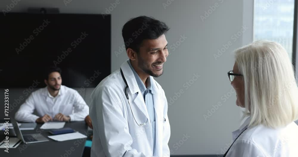 Positive young Indian doctor in uniform giving handshake to medical specialist woman with respect, gratitude, thanking for consultation, expertise, help, smiling. Hospital staff meeting in boardroom Positive young Indian doctor in uniform giving handshake to medical specialist woman with respect, gratitude, thanking for consultation, expertise, help, smiling. Hospital staff meeting in boardroom