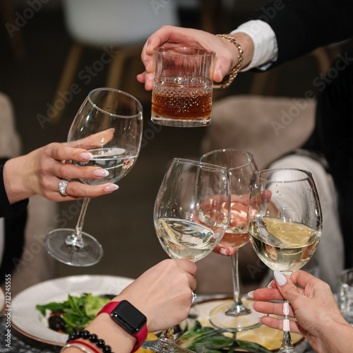 Cheers. Young friends clinking glasses and drinking. People celebrate and raise glasses of wine for toast. Group of men and women cheering with champagne. Happy new Year 2025 and holidays. Closeup.
