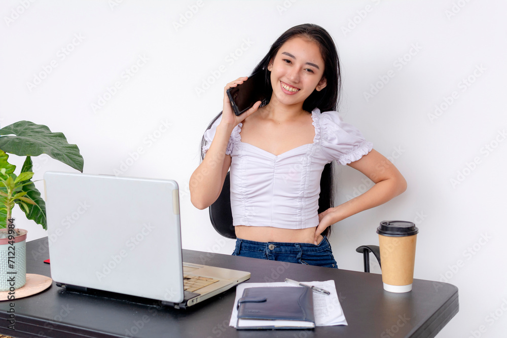 Young smiling female student using a mobile phone while standing by her desk with a laptop, coffee cup, and notes. Young smiling female student using a mobile phone while standing by her desk with a laptop, coffee cup, and notes.