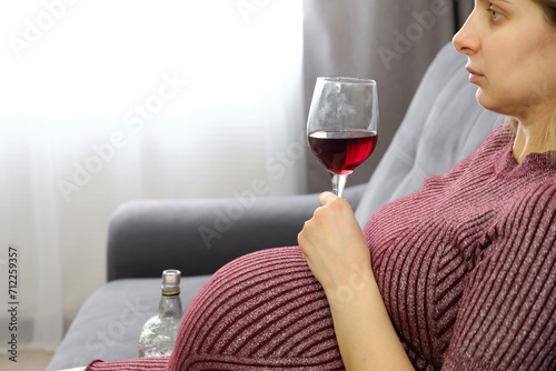 Depressed pregnant woman deciding whether or not to drink a glass of wine while sitting on the sofa. Caucasian expectant mother with bad and unhealthy habits