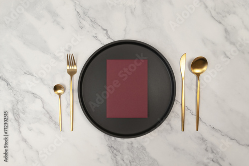 Top view of gold cutlery and black plate on white marble background. Table setting menu, empty paper blank flat lay. Fork, knife, spoon, dessert spoon, plate and blank. Copy space.