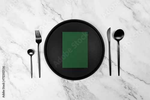 Top view of black cutlery and black plate on white marble background. Green blank, card flat lay. Copy space.