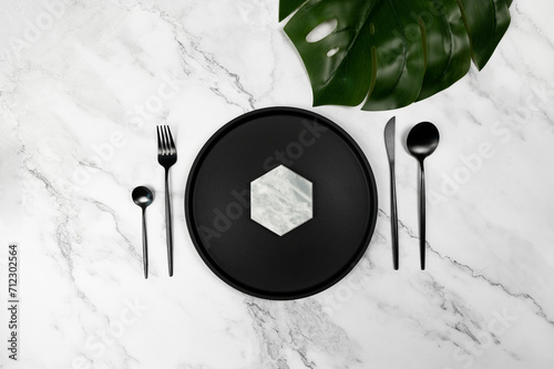Top view of black plate and black cutlery on white marble background. Luxury table setting flat lay. Fork, knife, spoon, dessert spoon and plate. Monstera tropical leaf, copy space.
