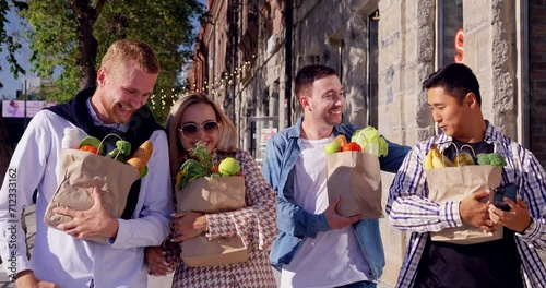 Group of four smiling friends together walk along street carrying bags with fresh food in sunny day. Shoppers happy with goods buys and groceries purchases photo