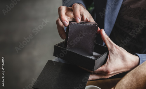 The man's hands take out a dark brown leather wallet from a black gift box. An elegant accessory symbolizes style, efficiency and attention to detail