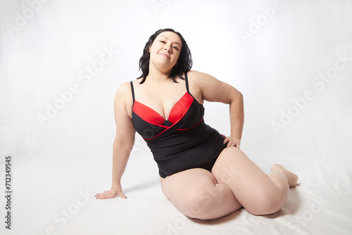 Portrait of attractive dreamy thick woman in red black swimsuit posing on white background. Body positive, photo shoot, selfie. Funny plus size model