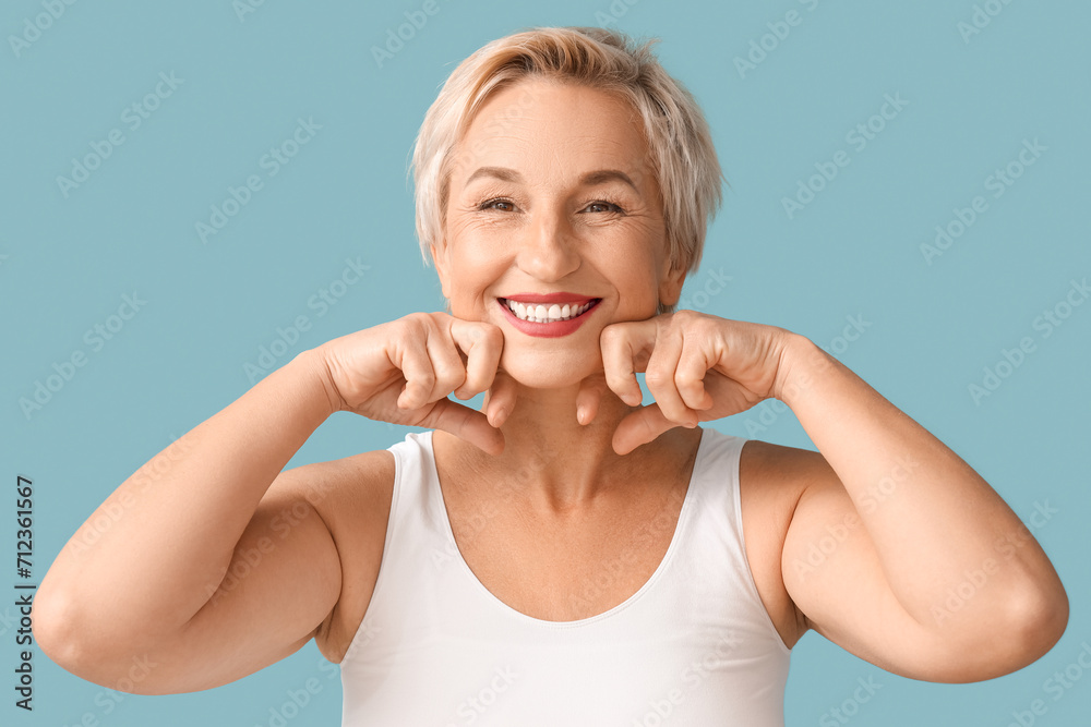 Fototapeta premium Beautiful mature woman doing face building exercise on blue background, closeup