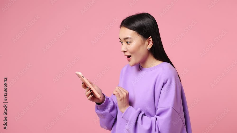 Young woman of Asian ethnicity wear purple sweatshirt hold use mobile cell phone typing browsing swipe chat send sms shopping online order delivery to home book tour isolated on plain pink background Young woman of Asian ethnicity wear purple sweatshirt hold use mobile cell phone typing browsing swipe chat send sms shopping online order delivery to home book tour isolated on plain pink background