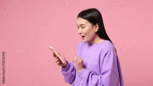 Young woman of Asian ethnicity wear purple sweatshirt hold use mobile cell phone typing browsing swipe chat send sms shopping online order delivery to home book tour isolated on plain pink background photo