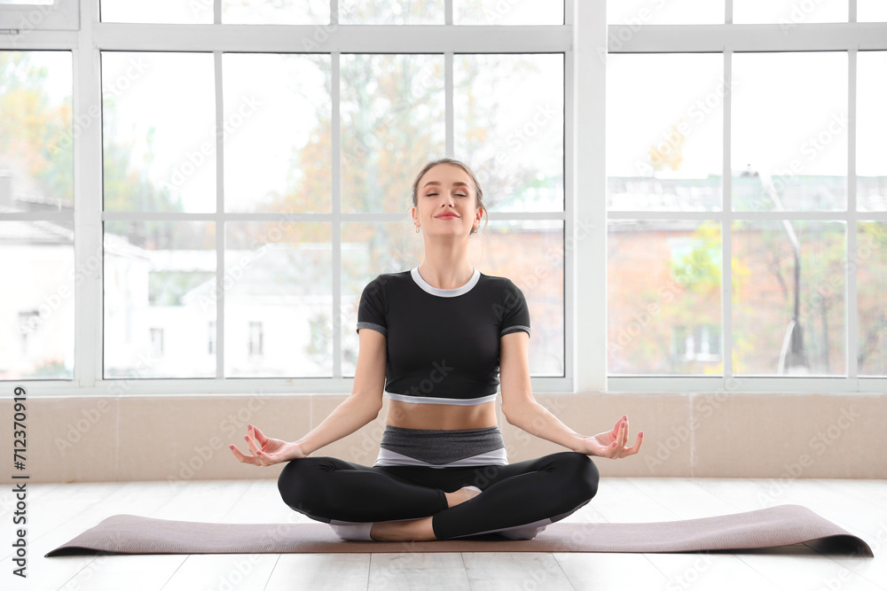 Sporty young woman meditating on yoga mat near window in gym
