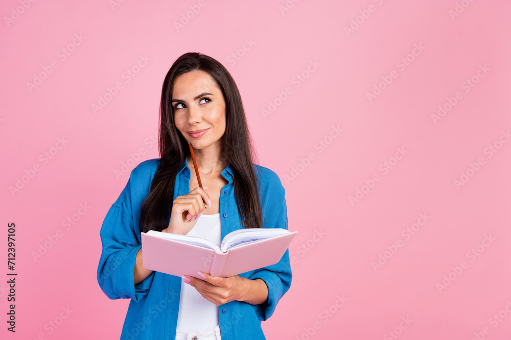 Photo of minded girl with straight hair dressed blue shirt hold notebook pencil on chin look empty space isolated on pink color background Photo of minded girl with straight hair dressed blue shirt hold notebook pencil on chin look empty space isolated on pink color background
