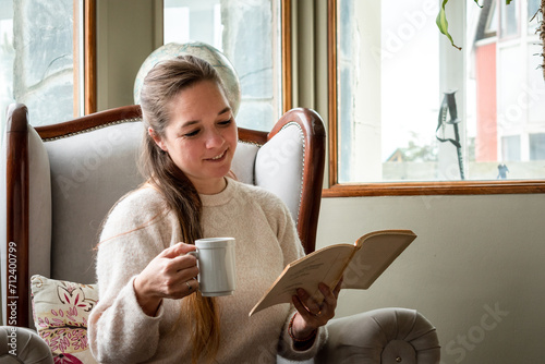 Mujer leyendo un libro mientras toma una taza de café, cómodamente en su sala de estar photo