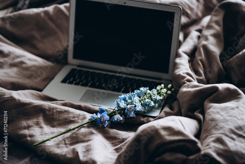 laptop in bed with gray sheets with blue delphinium flower. work from home romance at a distance. care for a loved one