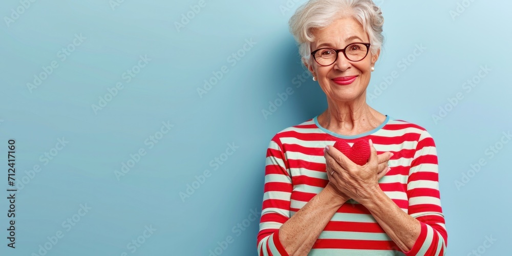 An elegant, fit granny with a charming smile, wearing glasses, holds a red apple in the kitchen. - obrazy, fototapety, plakaty 