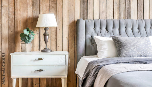 Bedside drawer nightstand and lamp near bed with grey fabric headboard against wood paneling wall. Farmhouse interior design of modern bedroom.