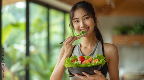Asian attractive sport woman holding salad bowl and eat vegetables. Beautiful sport girl in sportswear enjoy eat clean vegetables after exercise for health in house. Diet and Healthy food concept. 