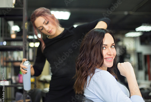 Young female hairdresser fixing hair of gorgeous woman with hairspray