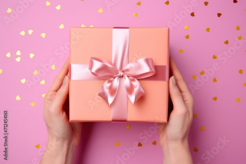 Hands of young woman holding beautiful gift box with pink ribbon on pink background with scattered confetti. Surprise gift in hands close-up. Congratulations on the holiday, March 8, Mother's Day © FoxTok