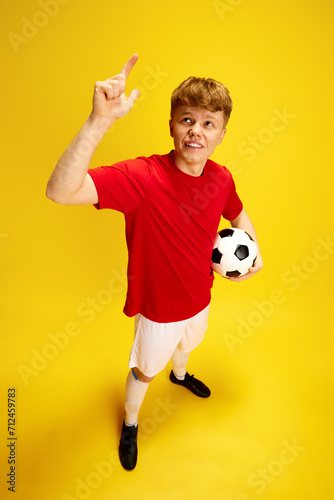 Fototapeta Naklejka Na Ścianę i Meble -  Top view portrait of young guy with freckles in football uniform, holding soccer ball against yellow studio background. Sport fan, betting. Concept of active lifestyle, youth, hobby and human emotions