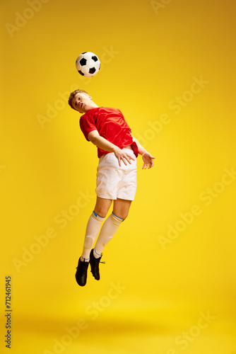 Full-length image of young man in uniform, soccer player, enthusiast in motion, training, hitting ball with head over yellow studio background. Concept of active lifestyle, youth, hobby and emotions