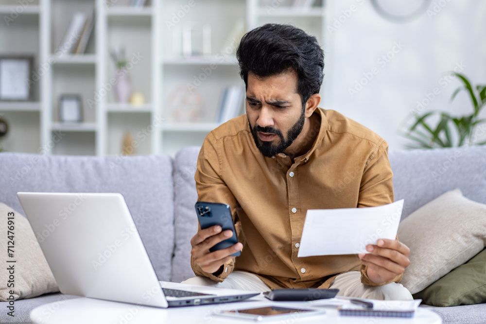 Indian man sitting on a sofa, looking at bills with concern while holding a phone with laptop and calculator nearby. Indian man sitting on a sofa, looking at bills with concern while holding a phone with laptop and calculator nearby.