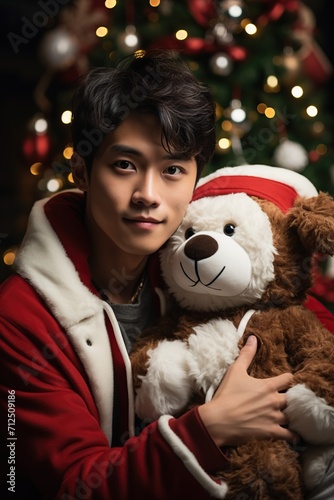 handsome and cute young guy sitting near the Christmas tree and hugging a teddy bear in a santa claus costume © Rafael