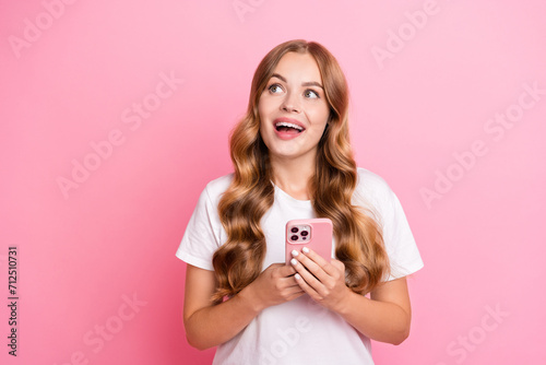 Portrait of ecstatic girl curly hair dressed white t-shirt holding smartphone look at offer empty space isolated on pink color background