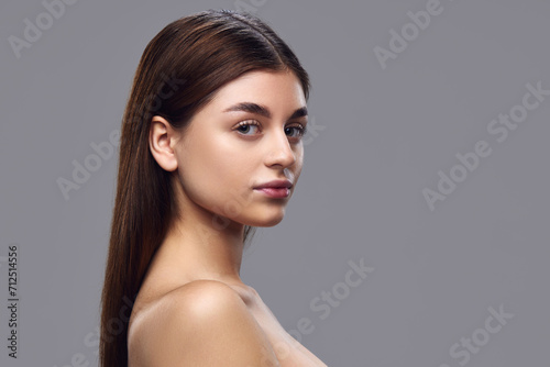 Portrait of young woman with clear skin, natural makeup, and neat hair looking over shoulder at camera against gray background. Concept of natural beauty, skin care products, nutritional supplements.