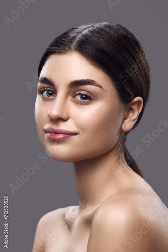 Portrait of young pretty brunette woman, female model looking at camera against grey studio background. Concept of makeup application highlighting fresh, everyday look.