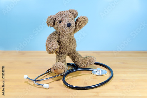 Little brown teddy bear standing on a stethoscope and waving, health care for children, pediatric medicine concept, blue background with copy space