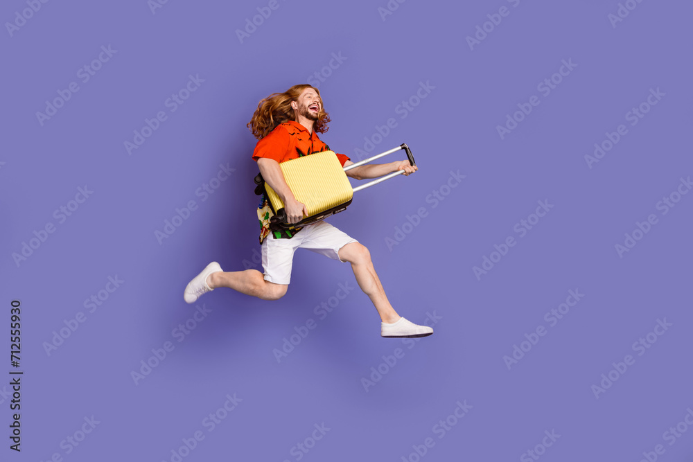 Fototapeta premium Full size photo of nice man wear print shirt jumping with suitcase hurry run to plane look empty space isolated on purple color background
