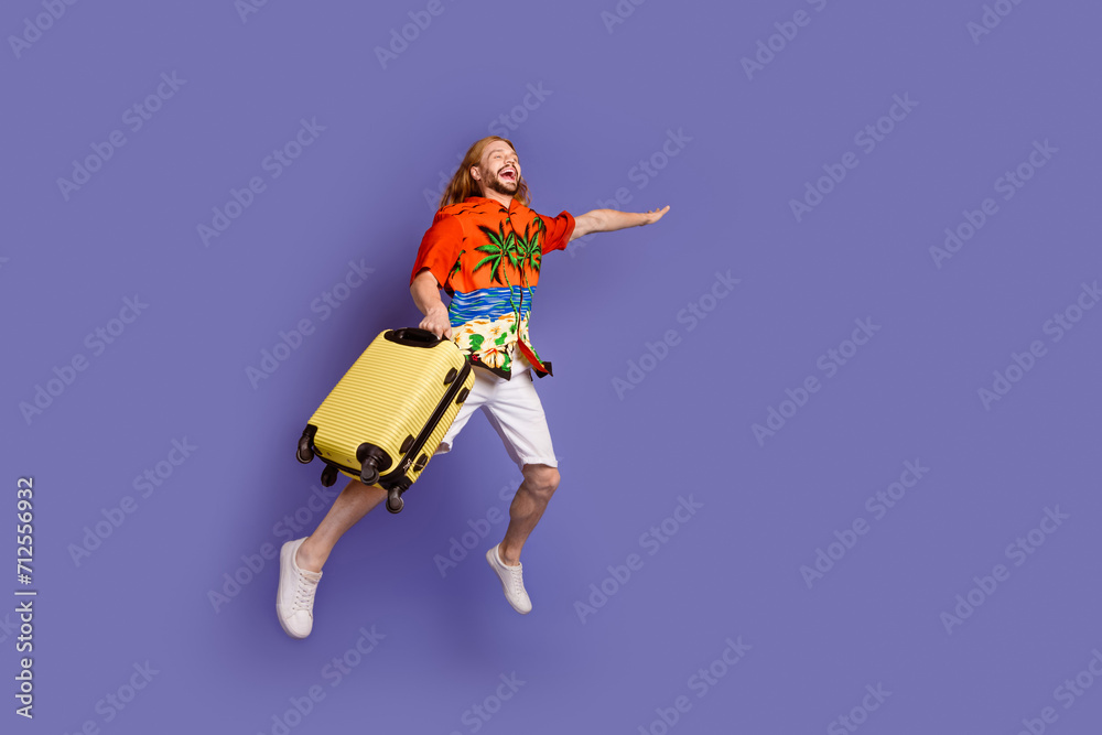 Full size photo of satisfied man wear print shirt white shorts jumping with suitcase look empty space isolated on purple color background Full size photo of satisfied man wear print shirt white shorts jumping with suitcase look empty space isolated on purple color background