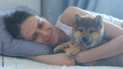 Happy young woman hugs cute puppy shiba inu dog lying on bed together at home interior. Close up. Young people, domestic animals, concept of happiness, friendship and love between an animal and human photo