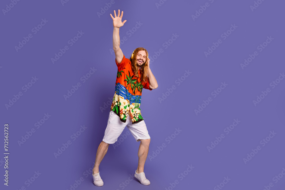 Full size photo of cool man wear print shirt white shorts touch headphones raising hand up say hi isolated on purple color background Full size photo of cool man wear print shirt white shorts touch headphones raising hand up say hi isolated on purple color background